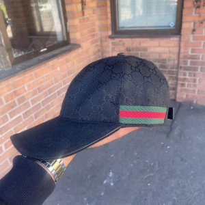 Svart keps från Gucci - Snygg svart keps från Gucci med diskret mönster och klassiska gröna och röda ränder på sidan. Kepsen har justerbart band för perfekt passform och är tillverkad i Italien. Perfekt för att ge en lyxig touch till din outfit.