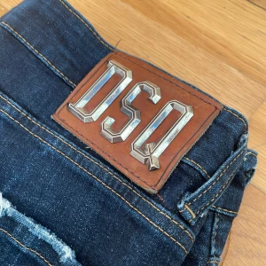 Mörkblå jeans från Dsquared2 - Snygga mörkblå jeans från Dsquared2  med slitna detaljer och en cool logga på baksidan. De har en klassisk femficksdesign och en knappgylf. Perfekta för en avslappnad stil.