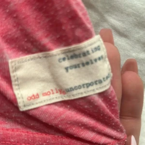 Rosa topp från Odd Molly - Säljer en rosa topp från Odd Molly med trycket 'Rise Above' och en ros på framsidan. Toppen har korta ärmar och en knytning vid halsen. Perfekt för en avslappnad stil.