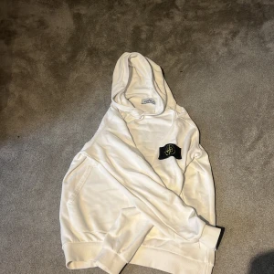 Vit hoodie från Stone Island - Säljer en stilren vit hoodie från Stone Island med deras ikoniska märke på ärmen. Perfekt för en avslappnad look. Hoodien har en klassisk passform och är tillverkad i mjukt material för extra komfort.