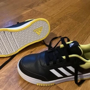 Svarta och gula sneakers från Adidas - Snygga svarta sneakers från Adidas med gula och vita detaljer. Skorna har en klassisk design med snörning och den ikoniska tre-randiga loggan på sidan. Perfekta för en sportig look.