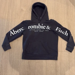 Amerikansk  Abercrombie & Fitch Hoodie - Säljer en svart hoodie från Abercrombie & Fitch med stor vit logga på bröstet och ärmarna. Hoodien har en klassisk känguruficka och justerbar huva med snören. Perfekt för en avslappnad stil.