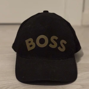 Svart keps från Boss - Snygg svart keps från Boss med justerbart spänne baktill. Kepsen har en stilren design med texten 'BOSS' i guld på framsidan. Perfekt för en casual look.
