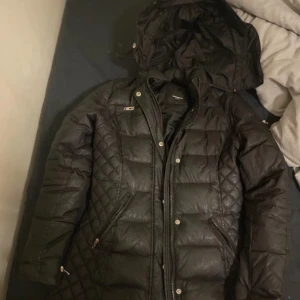 Svart dunjacka från Rockandblue - Snygg svart dunjacka från Rockandblue med quiltad design och avtagbar fuskpäls på huvan. Jackan har dragkedja och knappar framtill samt en liten ficka med dragkedja på ärmen. Perfekt för kalla dagar!