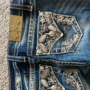 Blå jeans med broderade detaljer från Miss Me - Snygga blå jeans från Miss Me med unika broderade detaljer och paljetter på bakfickorna. Jeansen har en klassisk femficksdesign och en bekväm passform. Perfekta för att ge din outfit en extra touch av stil.