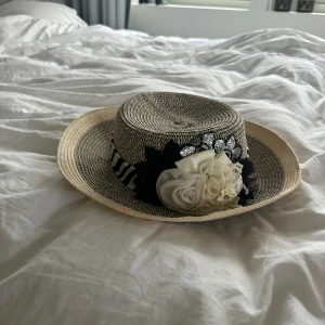Elegant stråhatt med blommor och paljetter - Snygg stråhatt med svartvitt mönster och en dekorativ randig band. Hatten har en stor vit blomma med pärlor och paljetter som ger en extra touch av elegans. Perfekt för sommarens alla tillfällen.