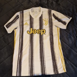 Juventus tröja från adidas - Säljer en vit och svart randig fotbollströja från Adidas med gulddetaljer. Tröjan har korta ärmar och en V-ringad krage. Den är märkt med 'Jeep' och har två stjärnor ovanför klubbmärket. Perfekt för fotbollsfans!