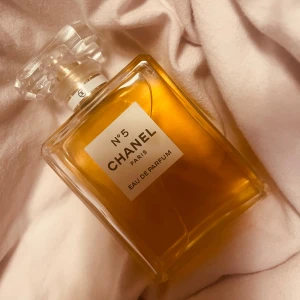 Chanel N°5 Eau de Parfum - Ikonisk Chanel N°5 Eau de Parfum i en elegant rektangulär glasflaska med en klassisk etikett. Parfymen har en gyllene färg och kommer med en stilren, fyrkantig kork. Perfekt för den som älskar tidlös elegans och lyx.