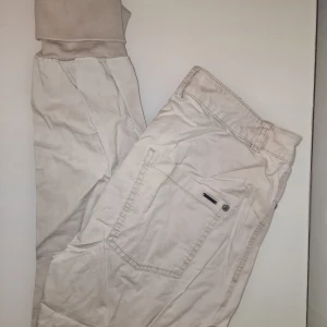 Beige byxor med knappar - Snygga beige byxor med flera knappar framtill. De har en avslappnad stil med ribbade muddar vid bensluten. Garderobsrensning så kika gärna på övriga auktioner.