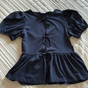 Marinblå (mörkblå) blus med knytband - Säljer en marinblå (mörkblå) blus med korta puffärmar och knytband framtill. Blusen har en volangdetalj vid midjan som ger en fin siluett. Perfekt för en stilren look. Den är öppen framtill med knytbandet! Rosett banden går inte att knyta upp utan det sitter fast på tröjan som en rosett, så det sitter så som det gör och går inte att ändra! 