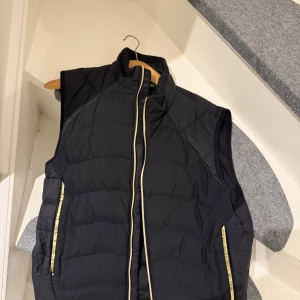 Svart dunväst med dragkedja - Snygg svart dunväst från Boss med dragkedja och gula detaljer. Västen har en quiltad design och är perfekt för kyligare dagar. Den har en hög krage och praktiska fickor med dragkedja. Nypris 3499, säljs för 599