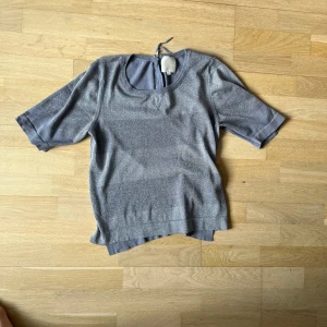 Glittrig grå topp - Snygg glittrig grå topp med rund hals och trekvartsärmar. Perfekt för en kväll ute eller en festlig tillställning. Toppen har en knytning i nacken för extra detalj.