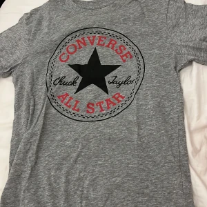 Grå t-shirt från Converse - Snygg grå t-shirt från Converse med det klassiska 'Chuck Taylor All Star' trycket i svart och rött. Perfekt för en avslappnad stil. T-shirten har en normal passform och korta ärmar❤️