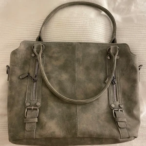 Grå axelväska med dragkedjor - Snygg grå axelväska med dubbla handtag och stilrena dragkedjedetaljer framtill. Väskan har en praktisk dragkedja baktill och ett rymligt innerfack med extra ficka. Perfekt för att bära med sig allt man behöver under dagen.