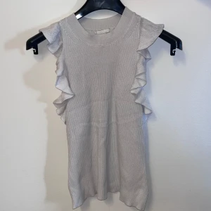 Beige ribbad topp med volangdetaljer från H&M - Säljer en stilren beige ribbad topp från H&M med volangdetaljer längs sidorna. Toppen är ärmlös och har en rund halsringning. Perfekt för en chic och bekväm look. Storleken går även på xs. 