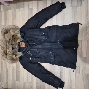 Svart parkajacka med pälsdetaljer - Säljer en svart parkajacka från Alpha Industries med pälsdetaljer på huvan. Jackan har flera fickor med knappar och dragkedja, samt en quiltad insida för extra värme. Perfekt för kalla dagar.