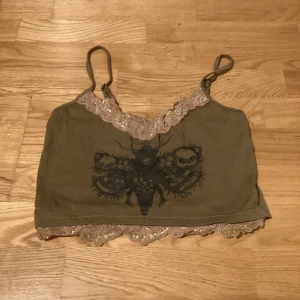 Olivgrön linnetopp med spetsdetaljer - Säljer en olivgrön linnetopp med smala axelband och spetsdetaljer i beige längs halsringningen och nederkanten. Toppen har ett tryck av en fjäril med dödskallar på framsidan. Via Swish.