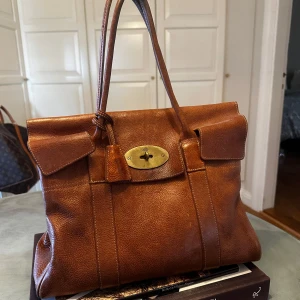 Bayswater Mulberry med metallspänne - Bayswater anses vara en ikon bland Mulberrys väskor. En tidlös och klassisk handväska som kombinerar det bästa av Mulberrys hantverk och eleganta stil. Både formen och designen lyfter fram den höga kvaliteten i lädret och matchas väl med Postman's Lock-stängningen. De justerbara bältesremmarna i sidorna ger möjlighet att anpassa väskans utrymme så att du alltid har en handväska anpassad för dina behov. Höjd: 26.5cm, bredd: 36cm, djup: 16.5cm, handtagshöjd: 15.5cm.  