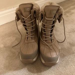 UGG adirondack - Snygga och varma beige vinterstövlar från UGG med snörning och mjukt pälsfoder. Perfekta för kalla dagar med bra grepp på sulan. Stövlarna har en stilren design med UGG-logga på sidan. Aldrig använda 