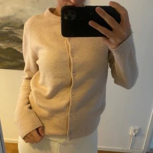 Beige kofta med knappar - Säljer en mysig beige/rosa kofta med knappar framtill. Koftan har långa ärmar och ribbstickade muddar vid ärmslut och nederkant. 