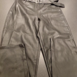 Svarta skinnbyxor med bälte - Snygga svarta skinnbyxor med medföljande bälte. Byxorna har en rak passform och dragkedja framtill. Perfekta för en stilren look.