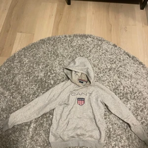 Grå hoodie från GANT - Säljer en stilren grå hoodie från GANT med broderad logotyp och emblem på bröstet. Tröjan har en klassisk design med huva och långa ärmar. Perfekt för en avslappnad stil.