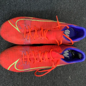 Röda Nike fotbollsskor - Snygga röda Nike fotbollsskor med gröna och blå detaljer. Skorna har snörning och en slitstark sula med dobbar för bra grepp på planen. Perfekta för fotbollsspelare som vill sticka ut.