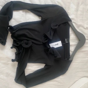 Svart volangtopp från Gina Tricot - Säljer en svart volangtopp från Gina Tricot i storlek XS. Toppen har långa ärmar och en croppad passform med volangdetaljer längs kanterna. Perfekt för en trendig look!