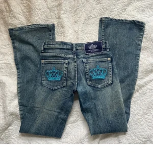 Blå jeans med broderade kronor - Snygga blå jeans från Victoria Bechem med unika turkosa broderade kronor på bakfickorna. De har en bootcut-stil och är perfekta för en avslappnad look. Jeansen har en klassisk femficksdesign och knappar med märkets logga. Det har en liten skråma på innersidan men inget som man tänker på när dom är på. 