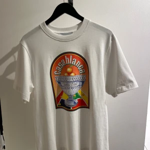 Vit t-shirt från Casablanca - Säljer en helt ny vit tshirt från Casablanca med ett coolt tryck på framsidan. T-shirten har korta ärmar och är perfekt för en avslappnad stil. Materialet känns mjukt och skönt mot huden. Passar perfekt till både jeans och shorts!