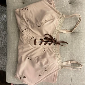 Beige korsettopp från Chemise - Y2k tröja, från 2000 talet. Säljer en superfin beige korsettopp från Chemise. Den har vackra spetsdetaljer och broderade blommor. Toppen har snörning framtill och justerbara axelband för perfekt passform. Storlek S