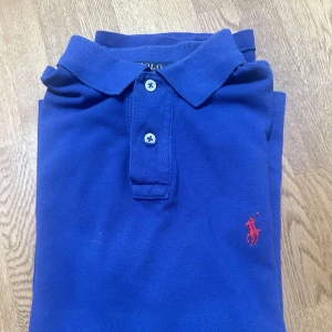 Blå pikétröja från Ralph Lauren - Säljer en snygg blå pikétröja från Ralph Lauren i storlek M. Tröjan har en klassisk krage med två knappar och det ikoniska röda Ralph Lauren-logotypen broderad på bröstet. Perfekt för både vardag och lite finare tillfällen!
