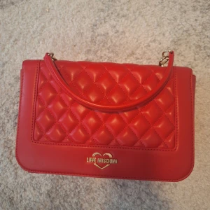 Helt ny Love Moschino väska - Väskan köptes för ca 3 år sedan för 2600kr men säljer för 2000kr. Pris kan diskuteras vid snabb affär. Väskan har alldrig blivit använd. Axelband och tygpåse tillhörande väskan medföljer. Längd: 30cm Höjd: 18cm