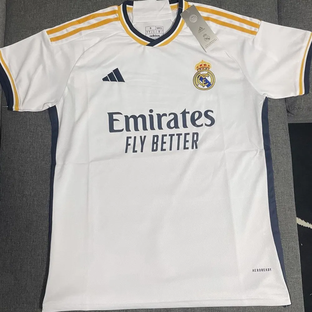 Säljer en snygg vit Real Madrid fotbollströja från Adidas. Den har korta ärmar och coola gula och svarta detaljer på axlarna. Perfekt för alla fotbollsfans som vill visa sitt stöd för laget! Tröjan är i nyskick och har fortfarande taggen kvar.. T-shirts.