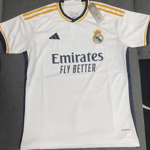 Vit Real Madrid fotbollströja - Säljer en snygg vit Real Madrid fotbollströja från Adidas. Den har korta ärmar och coola gula och svarta detaljer på axlarna. Perfekt för alla fotbollsfans som vill visa sitt stöd för laget! Tröjan är i nyskick och har fortfarande taggen kvar.