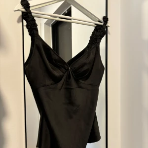 Svart satinlinne med rynkade axelband - Snyggt svart linne i satin från adoore med rynkade axelband. Perfekt för en utekväll eller fest. Linnet har en liten detalj framtill som ger det en extra snygg touch.😍