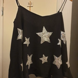 Svart linne med stjärnor från Hollister - Säljer ett snyggt svart linne från Hollister med tunna axelband. Toppen är dekorerad med glittriga stjärnor i olika storlekar, vilket ger en festlig känsla. Perfekt för en utekväll eller fest!