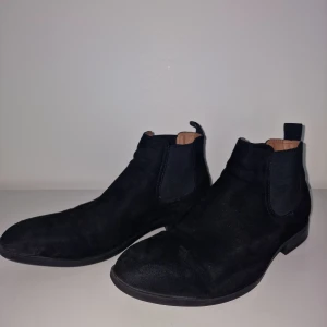 Svarta Chelsea boots i mocka - Säljer ett par stilrena svarta Chelsea boots i mocka. De har en klassisk design med elastiska paneler på sidorna för enkel på- och avtagning. Perfekta för både vardag och fest, och de ger en snygg touch till vilken outfit som helst. Använda men i bra skick!