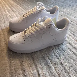 Säljer ett par klassiska vita Nike Air Force sneakers i nyskick. De har en stilren design med snörning och är perfekta för både vardag och fest. Skorna är i skinn och har en bekväm passform. Perfekta för alla säsonger!