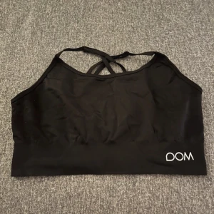 Svart sporttopp från DOM - Säljer en svart sporttopp från DOM med korsade band i ryggen. Perfekt för träning och ger bra stöd. Toppen är i ett mjukt och stretchigt material för maximal komfort.