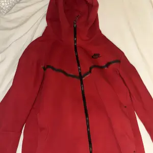 Säljer en snygg röd hoodie från Nike i storlek M. Den har 2 dragkedjor. Perfekt för både träning och vardag. Hoodien har långa ärmar och en stilren design med svarta detaljer. Men är sönder vid en av fickorna men går enkelt fixa om man kan sy!