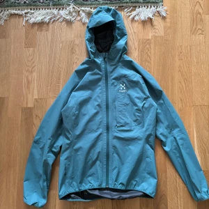 Blå regnjacka från Haglöfs - Säljer en snygg blå regnjacka från Haglöfs i storlek M för dam. Jackan är tillverkad i Gore-Tex-material vilket gör den perfekt för regniga dagar. Den har en praktisk huva och en dragkedja framtill. Perfekt för höst- och vårsäsongen när vädret är lite osäkert.