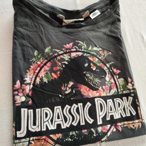 Svart Jurassic Park t-shirt - Säljer en svart t-shirt med coolt Jurassic Park-tryck. T-shirten har ett färgglatt blommönster runt dinosaurien och texten. Perfekt för filmälskare och vardagsbruk. Den är i bomull och har korta ärmar. Skön och stilren!