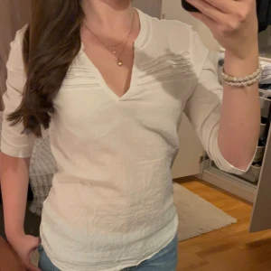 Vit topp från Caroll - Säljer en stilren vit topp från Caroll i storlek M. Den har en v-ringad design med fina detaljer vid axlarna. Perfekt för både vardag och festliga tillfällen. Toppen är i nyskick och gjord av ett mjukt och bekvämt material.