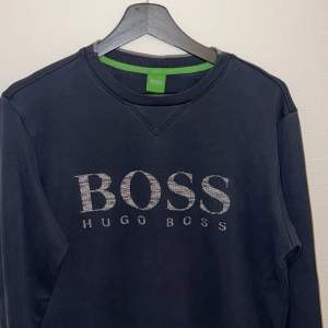 Säljer en snygg mörkblå sweatshirt från Hugo Boss. Tröjan har en klassisk design med BOSS-loggan i silver framtill. Den är i storlek M och passar perfekt för en casual look. Materialet känns mjukt och bekvämt, perfekt för både höst och vår. Perfekt för dig som vill ha en stilren och bekväm tröja i garderoben!