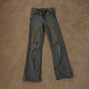 Grå jeans med slitningar - Säljer ett par grå jeans med coola slitningar på knäna. De har en klassisk femficksdesign och en knappgylf. Perfekta för en avslappnad och trendig look!
