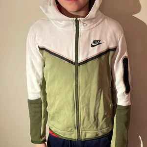 Grön och vit huvtröja från Nike - Säljer en snygg grön och vit huvtröja från Nike i storlek M. Tröjan har en dragkedja framtill och en praktisk ficka på ärmen. Perfekt för höst och vår, med en sportig stil och bekväm passform. Passar både till träning och vardag.