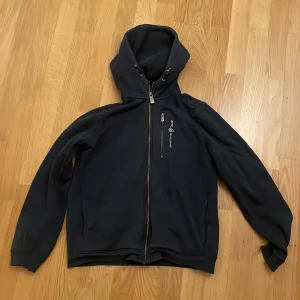 Mörkblå hoodie från Sail Racing - Säljer en mörkblå hoodie från Sail Racing i bra skick. Den har en dragkedja framtill och en mysig huva. Perfekt för höst och vinter. Det finns ett litet märke på ärmen, men inget som stör funktionen. 