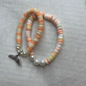 Färgglatt armband med pärlor och berlock - Säljer ett superfint armband med färgglada platta pärlor i orange, beige och rosa nyanser. Armbandet har också vita pärlor och en söt silverfärgad berlock i form av en valfena. Perfekt för sommarens alla äventyr! 🐋✨
