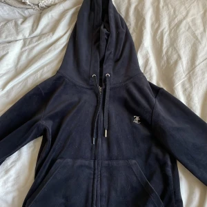 Svart hoodie från Juicy Couture - Säljer en svart Juicy Couture hoodie i velour med dragkedja och huva. Den har snygga strassdetaljer på ryggen som bildar texten 'Juicy'. Loggan har saknats sedan jag köpte den ny, en metalsak saknas även på ena bandet, har det hemma dock så kan limma på det om det önskas 